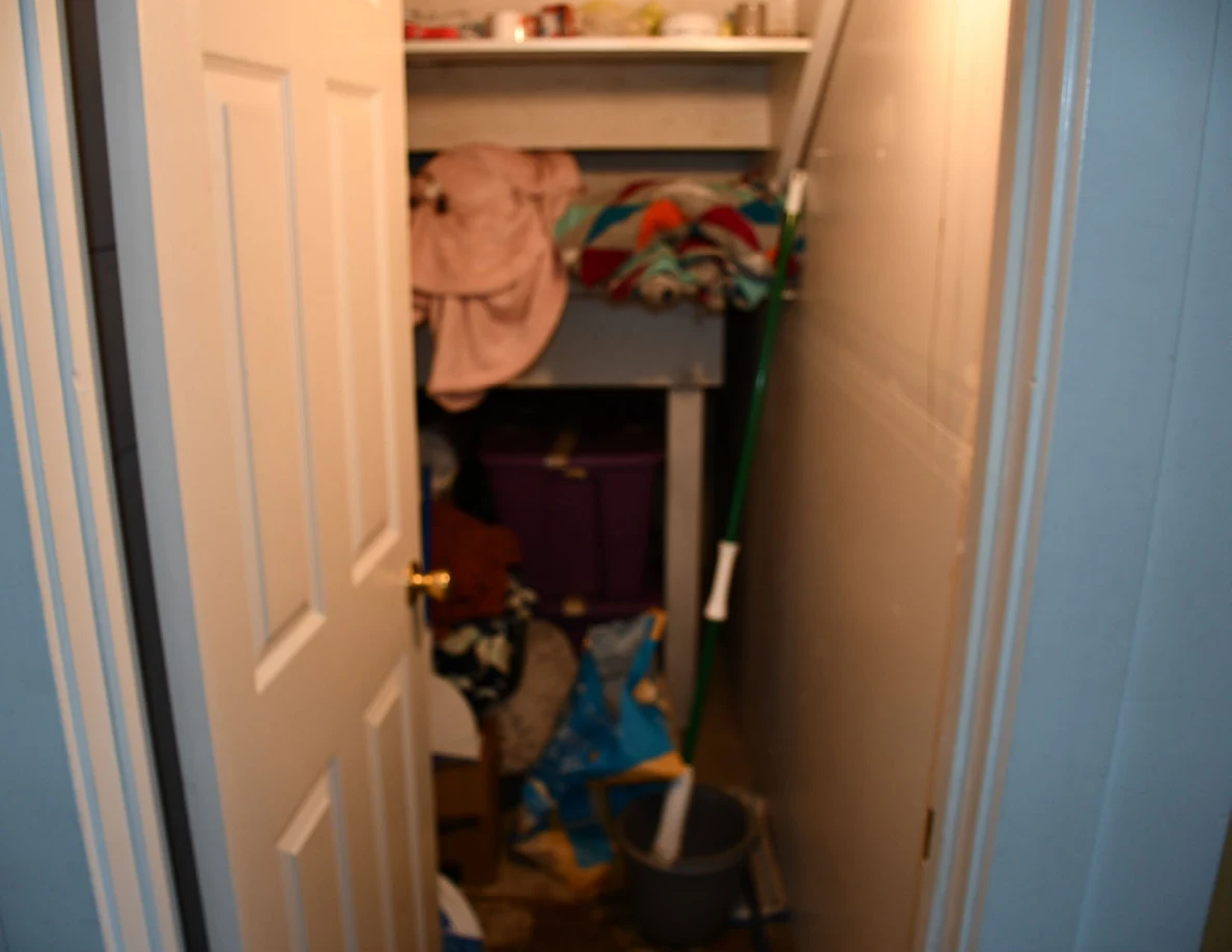 Open storage closet containing shelves, towels, a purple bin, bags and a mop bucket.
