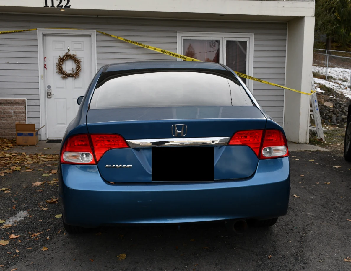 Rear of a blue Honda sedan outside a gray house marked 1122, with yellow tape and a blacked-out license plate.