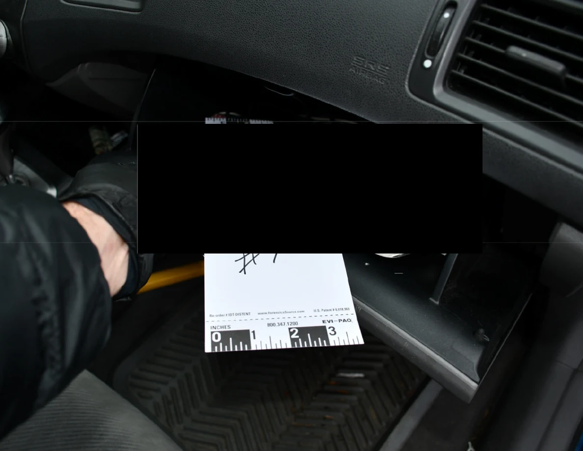 Gloved hand near an open glove compartment and measuring marker 7, with a large black redaction over the contents.