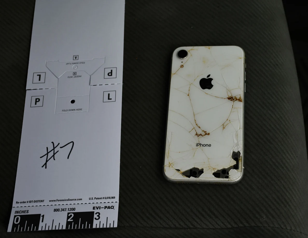 White iPhone with a cracked and chipped back on a gray vehicle seat beside measuring marker 7.