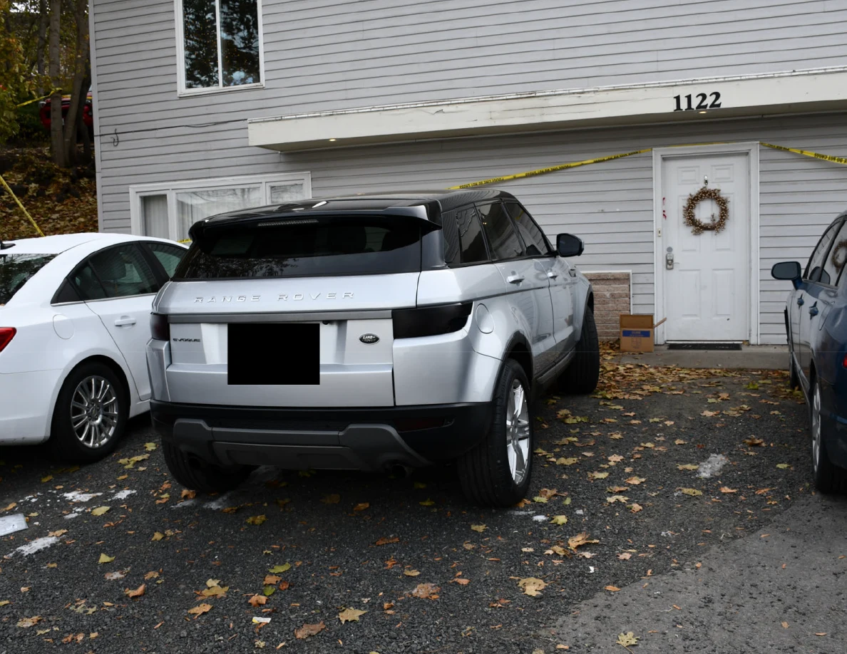 Rear of a silver Range Rover outside a gray house, with a blacked-out license plate and yellow tape.