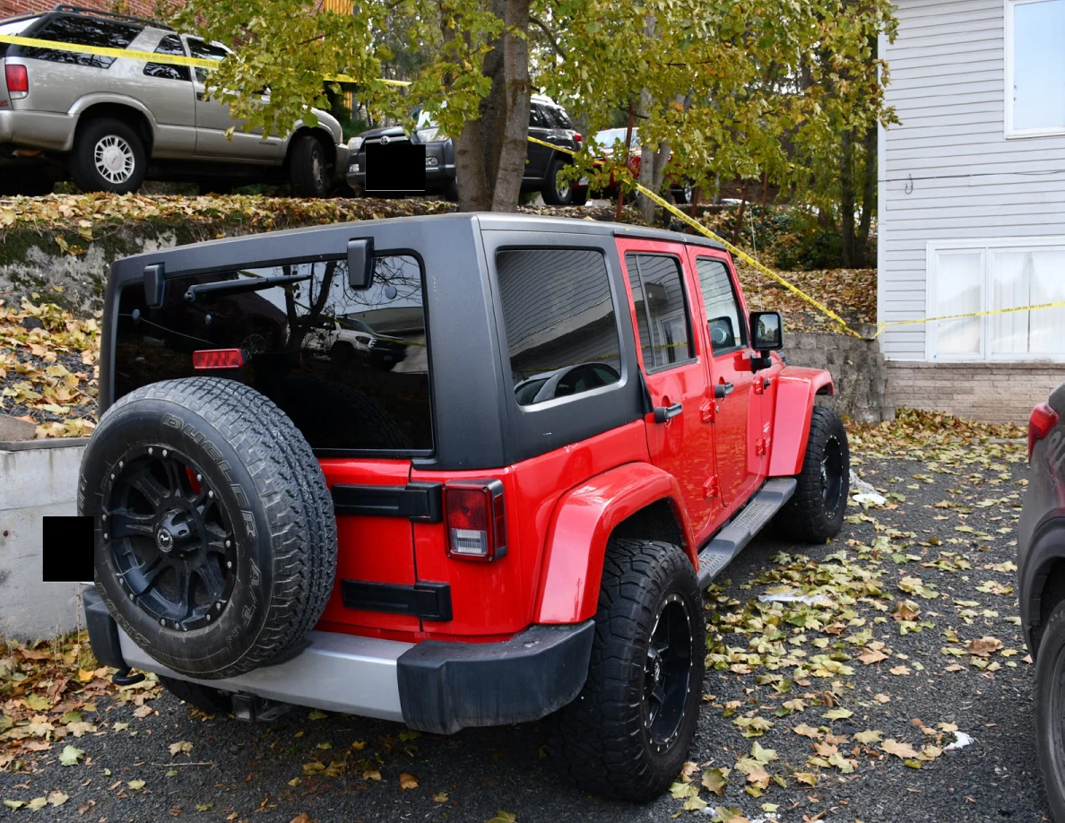 Rear and side of a red Jeep with a rear-mounted spare tire, parked among fallen leaves near yellow tape.