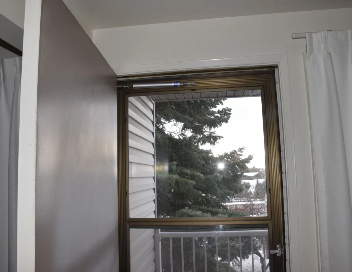Upper doorway and glass storm door beside curtains, with a tree and balcony railing outside.