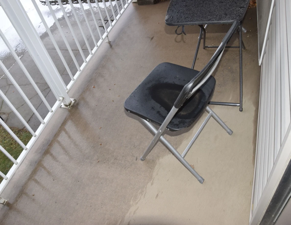Small balcony with a black folding chair, small table, white railing and wet floor.