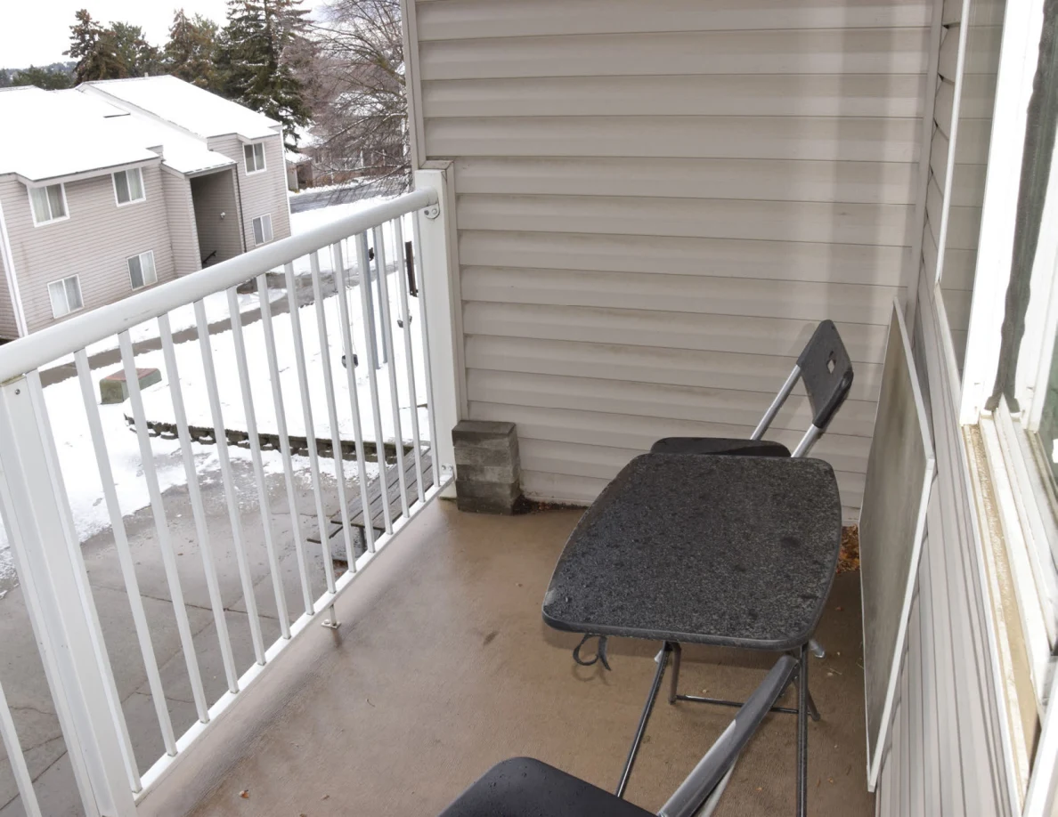 Balcony with a black folding chair and table beside pale siding and a white railing, overlooking snowy houses.