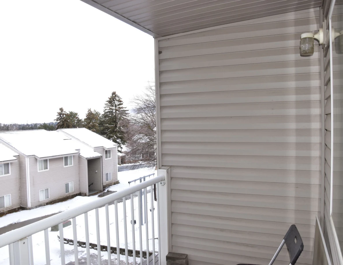 Balcony corner with pale siding, an exterior light and white railing, with snow-covered neighboring roofs beyond.