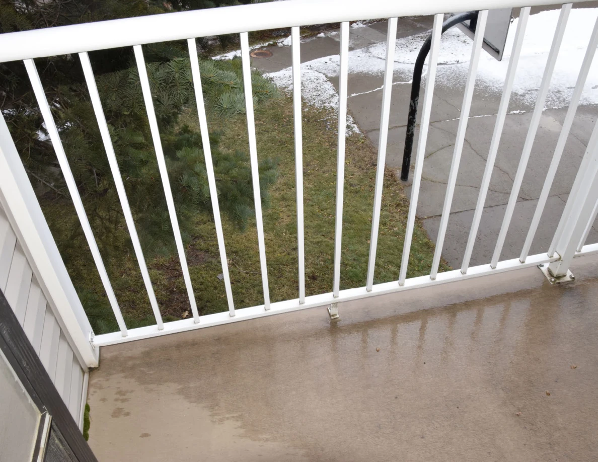 White balcony railing above a wet concrete floor, with a road and snow visible through the bars.