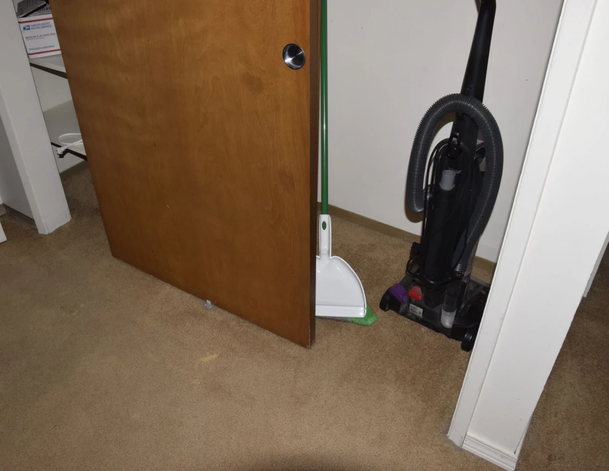 Partly open wooden sliding closet door revealing a vacuum cleaner and long-handled dustpan.