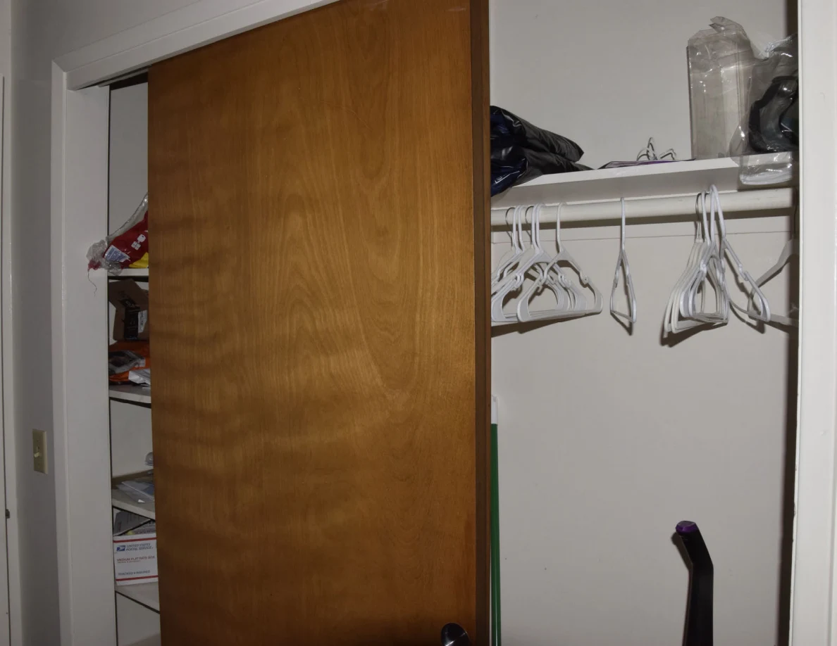 Closet with a wooden sliding door, empty white hangers and stored items on the upper shelf.