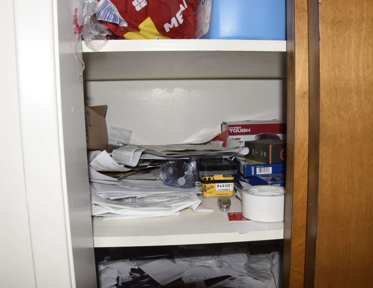 Storage shelf crowded with loose papers, small boxes, household supplies and a roll of tape.