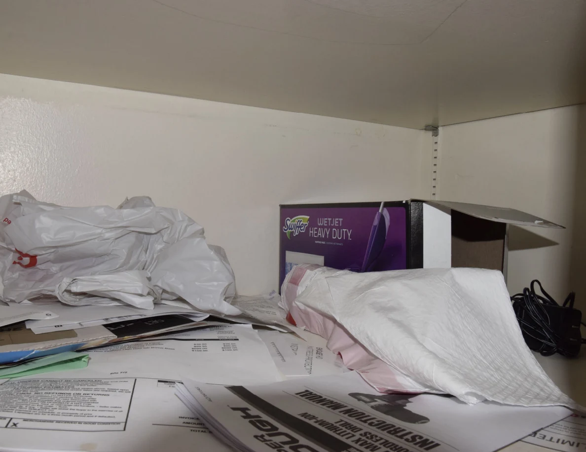 Storage shelf with papers, a folded white cloth, plastic bags and a purple cleaning-product box.