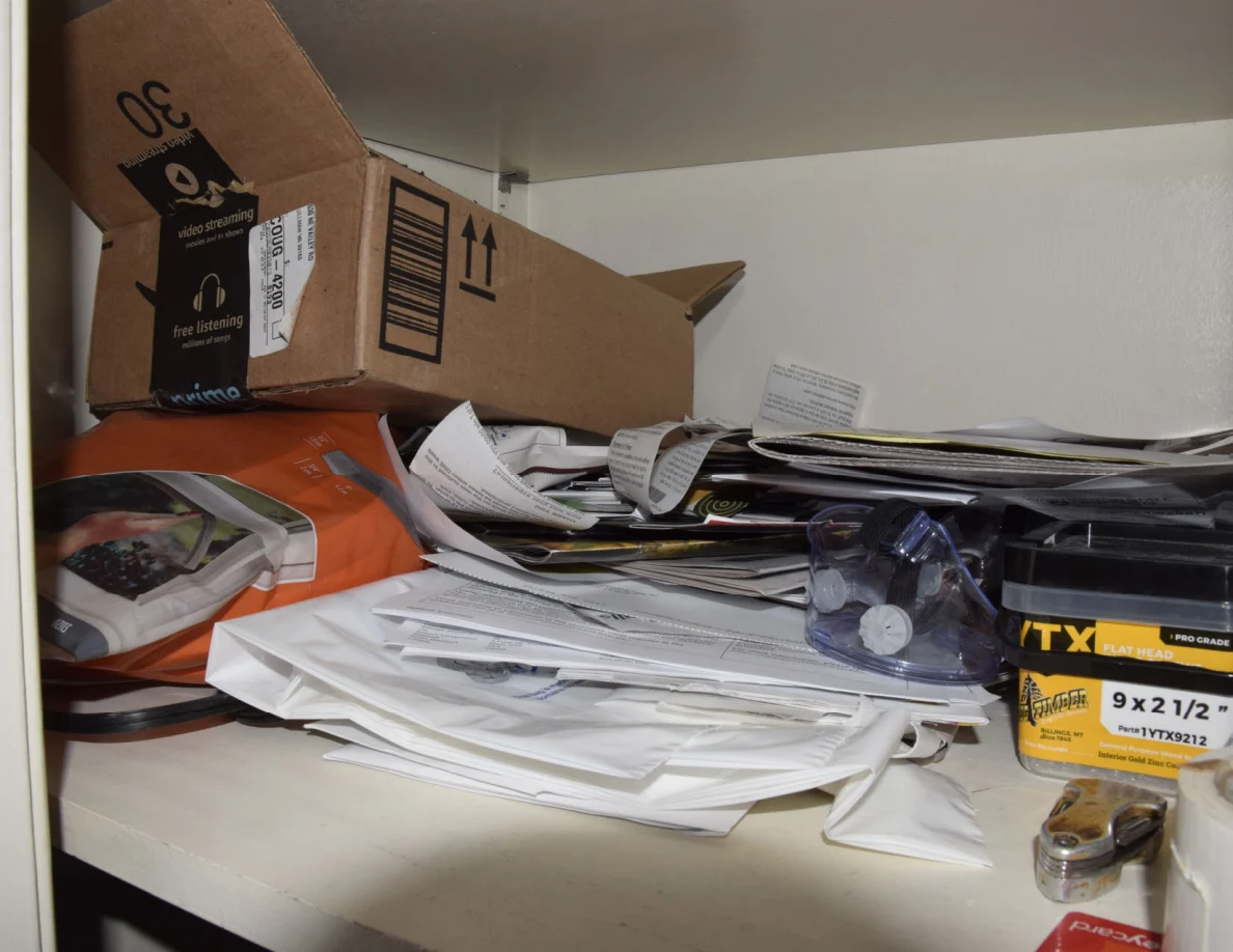 Shelf with a cardboard shipping box, stacks of papers and small packaged household supplies.