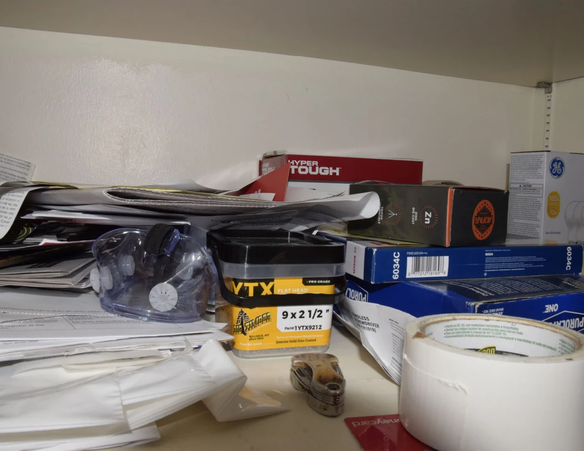 Storage shelf crowded with papers, hardware boxes, a tape measure and a roll of white tape.