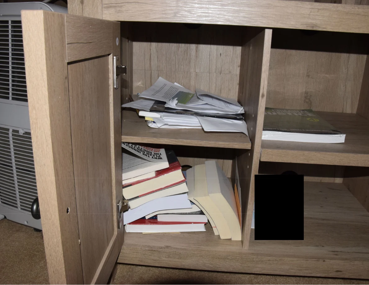 Open media-cabinet door revealing stacks of papers and books, with a black redaction on a lower shelf.