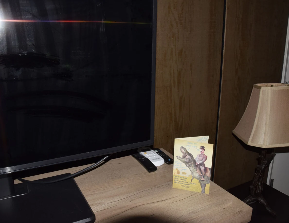 Television, remote controls and a yellow illustrated greeting card beside a table lamp.