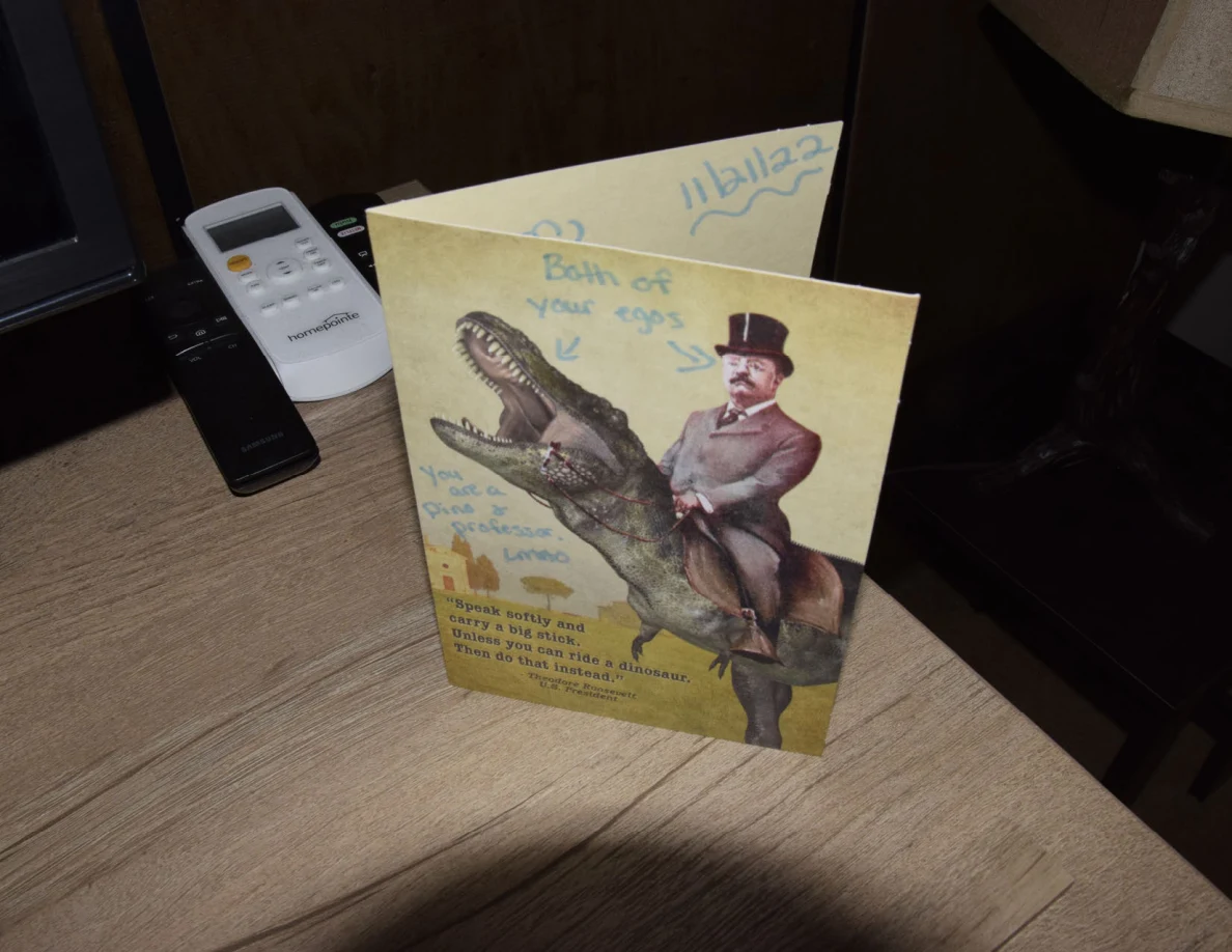 Open yellow greeting card depicting a rider on a dinosaur, with handwritten blue notes, beside remote controls.