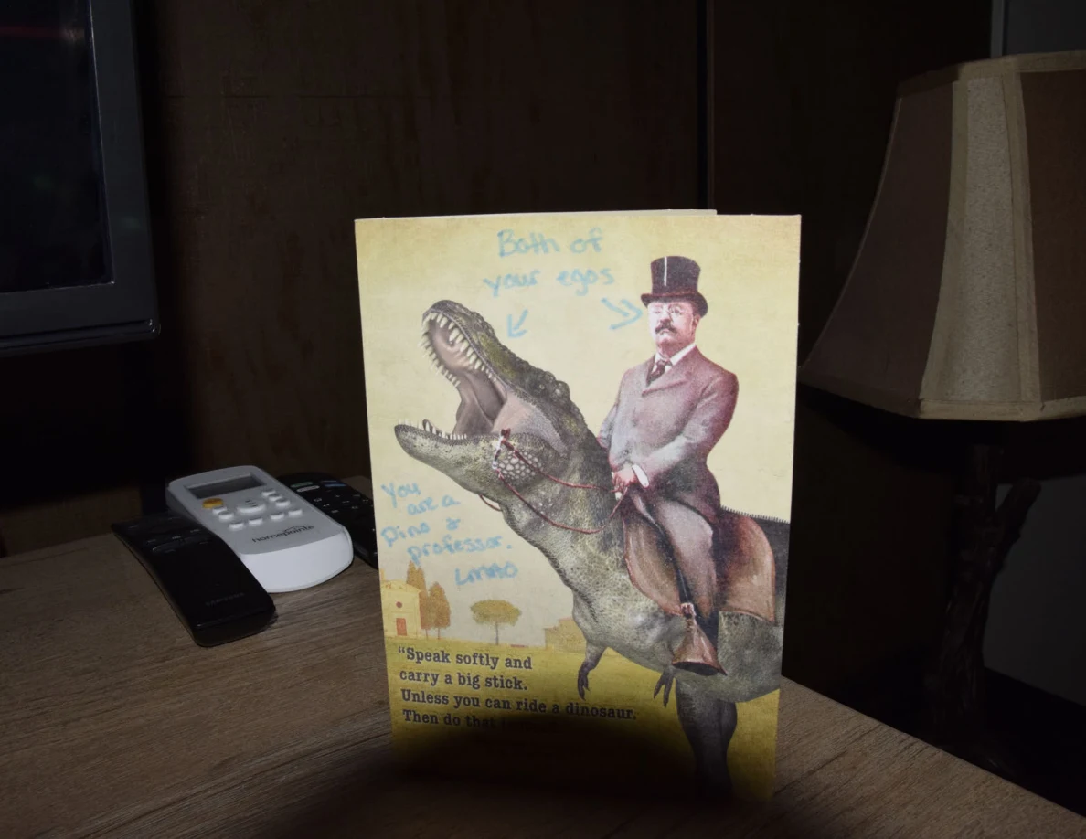 Front of a yellow dinosaur-rider greeting card with handwritten blue annotations, beside remote controls and a lamp.