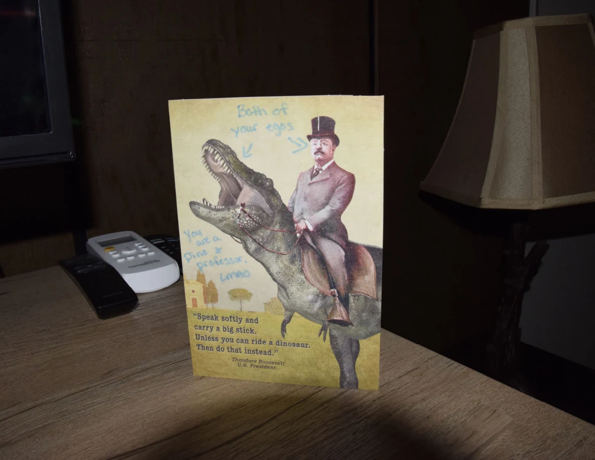 Upright yellow dinosaur-rider greeting card on a wooden cabinet beside a table lamp.