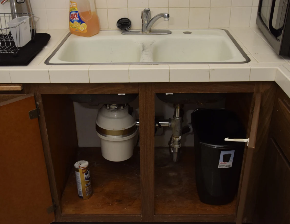 Kitchen double sink above an open cabinet showing a disposal unit, pipes, cleaner and black trash bin.