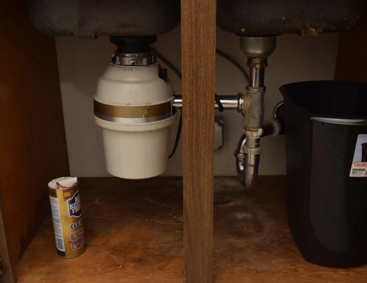 Under-sink cabinet with a disposal unit, drain pipes, cleanser canister and black bin.