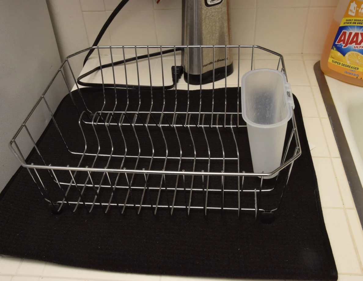 Empty metal dish rack on a black drying mat beside a grater and dish soap.