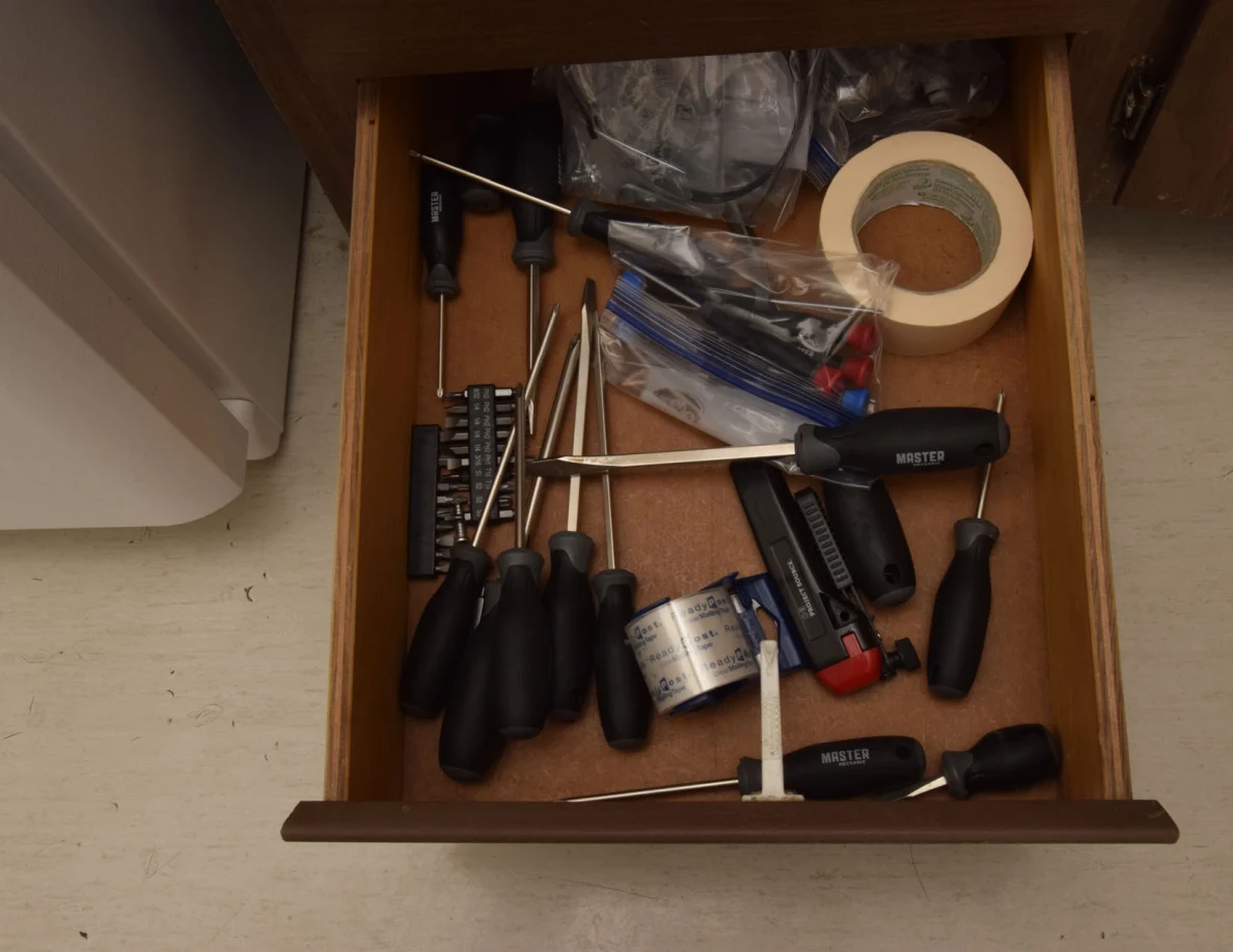 Open drawer containing screwdrivers, small tools, bags and a roll of masking tape.