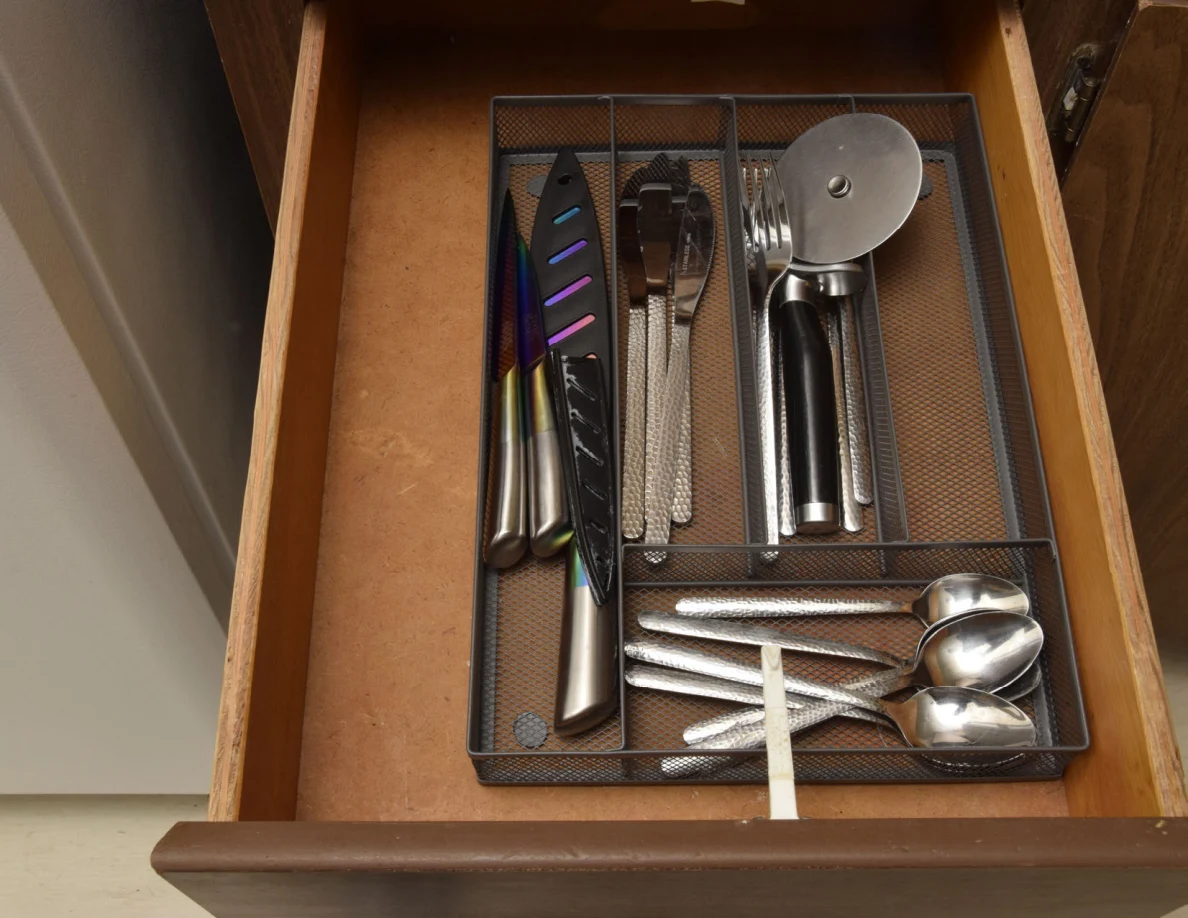 Open cutlery drawer with a mesh organizer, knives, forks, spoons and a pizza cutter.
