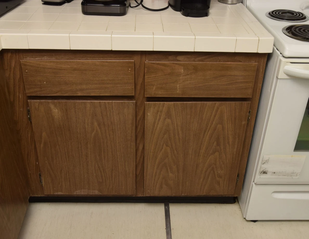 Closed wooden kitchen cabinet beneath a tiled countertop beside a white electric stove.