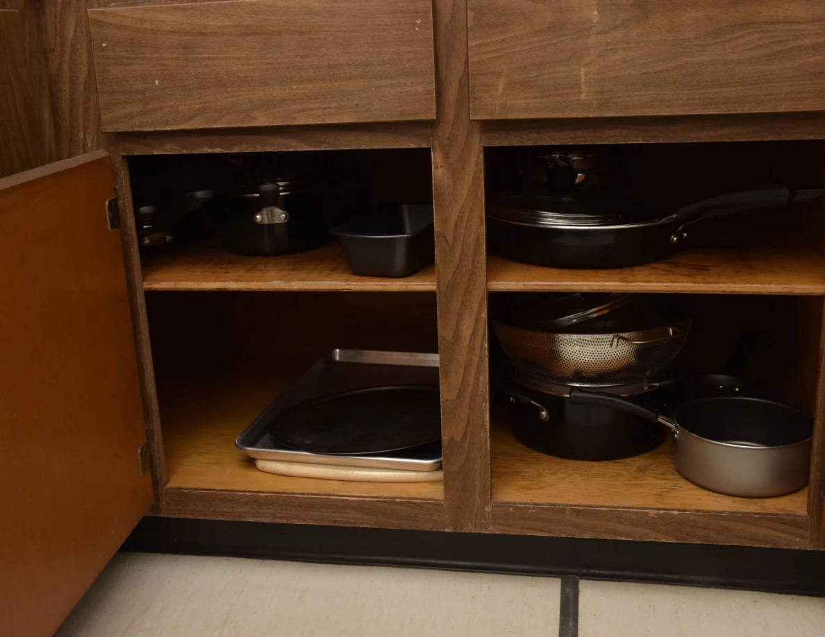 Open kitchen cabinet holding pots, frying pans, a colander and baking trays.