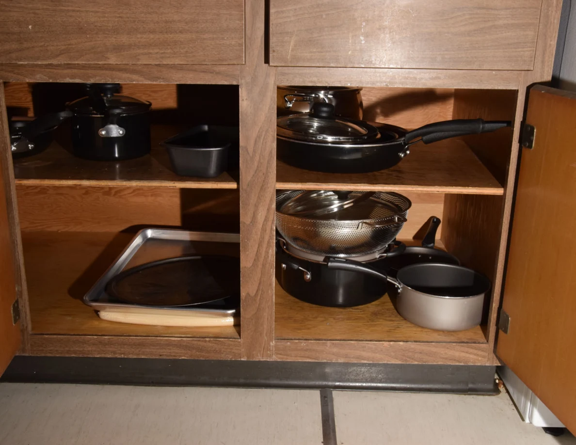 Open wooden cabinet with stacked pots, pans, lids, a colander and metal trays.