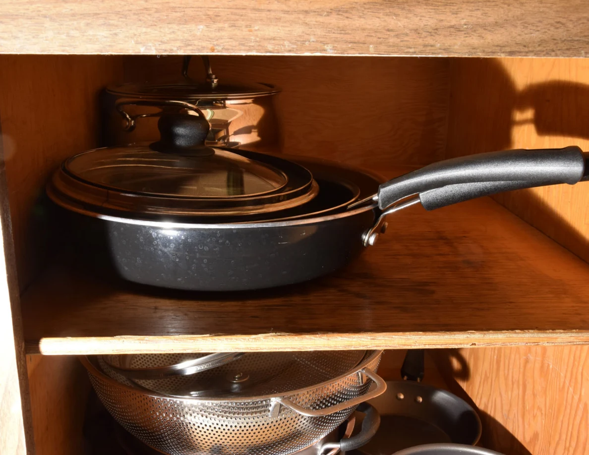 Lidded black frying pan on a cabinet shelf above stacked metal colanders.