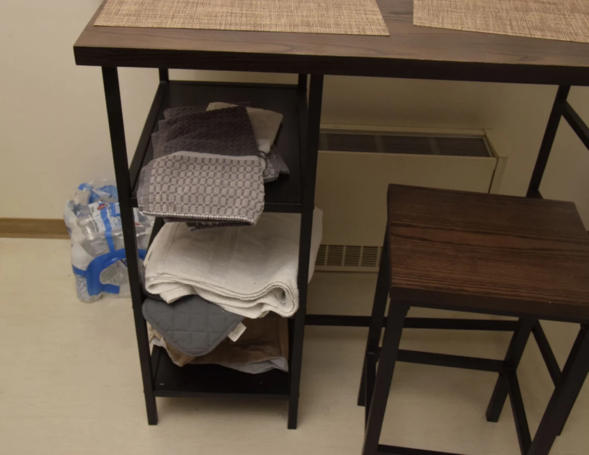 Small dark table and stool with placemats, folded towels on side shelves and bottled water on the floor.
