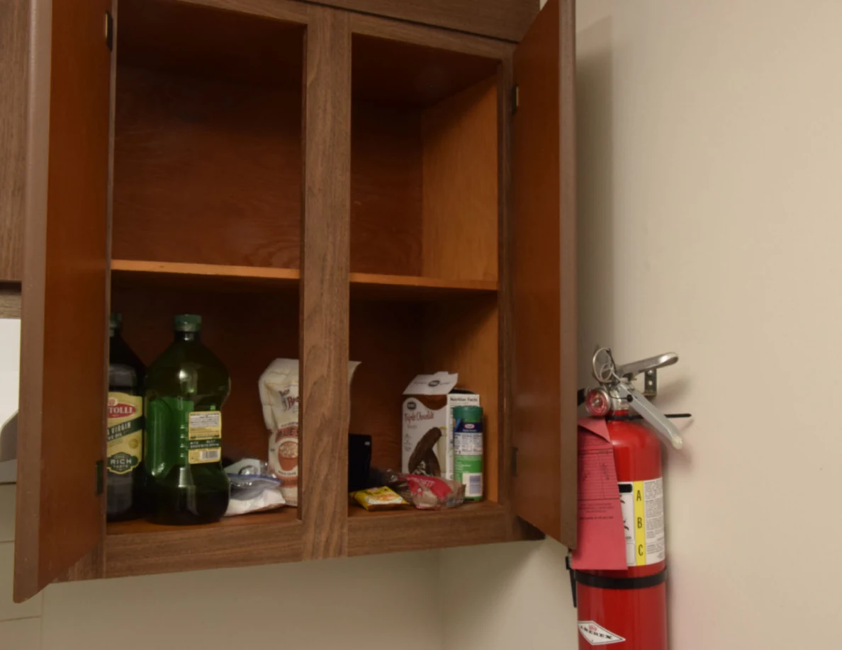 Open wooden food cupboard with bottles and dry goods on the bottom shelf, beside a fire extinguisher.