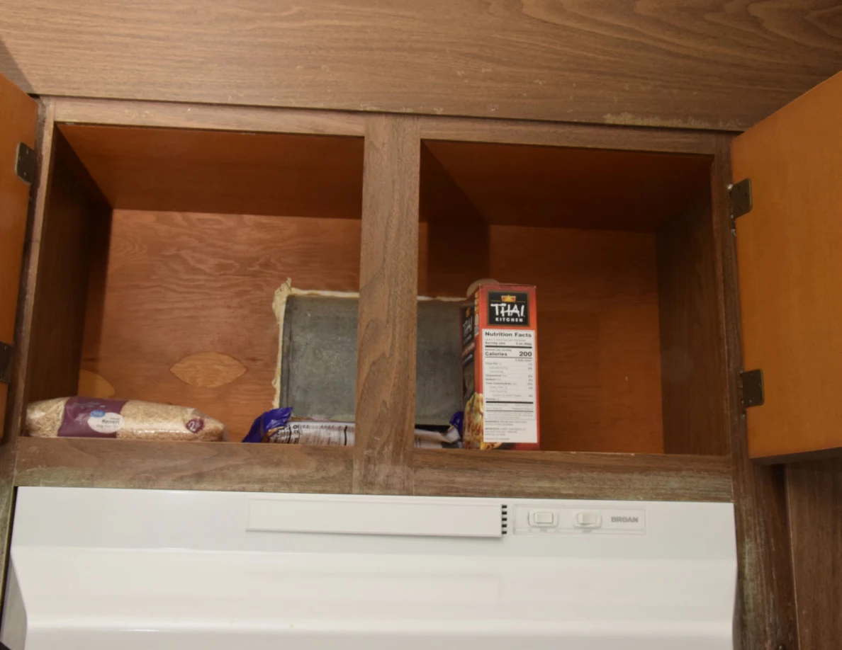 Open cupboard above a stove hood containing rice and boxed food beside a rectangular metal duct.