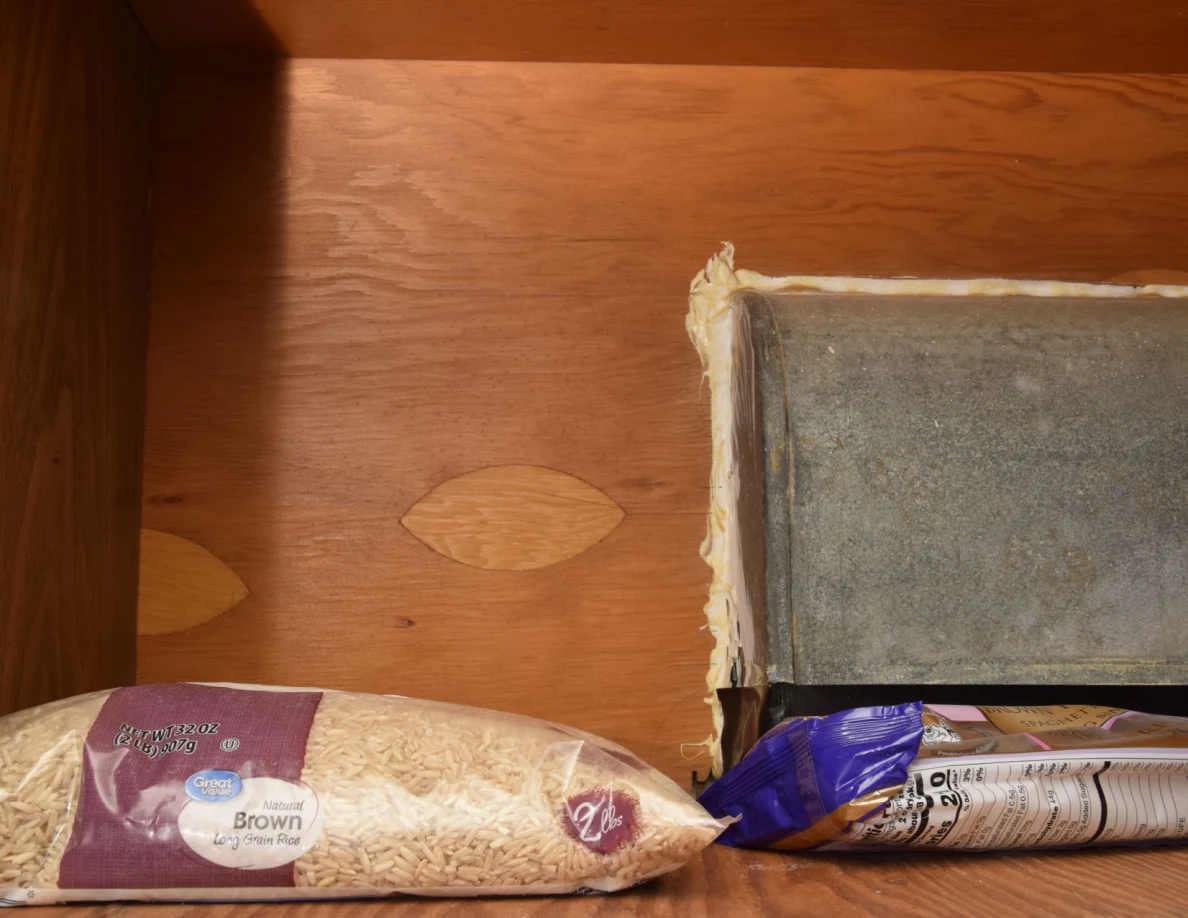 Bag of brown rice and food packets beside a metal duct inside a wooden cupboard.