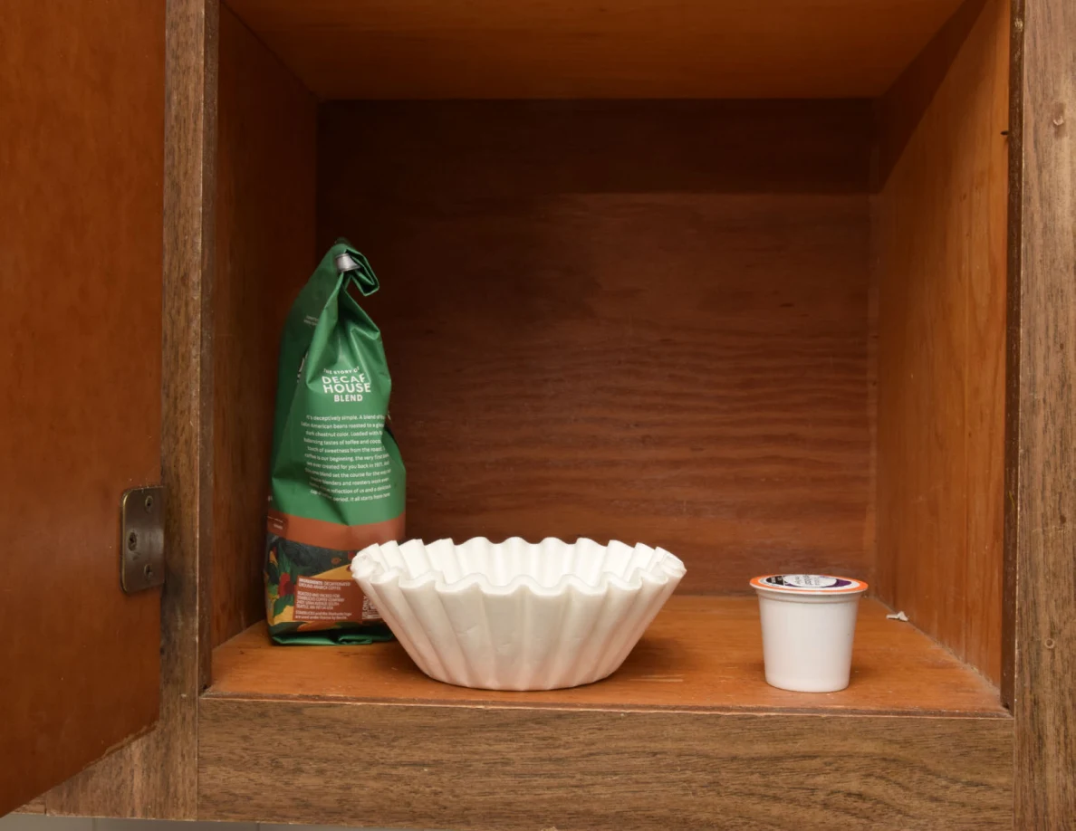 Coffee bag, fluted paper filters and a single-serve pod on a wooden cupboard shelf.