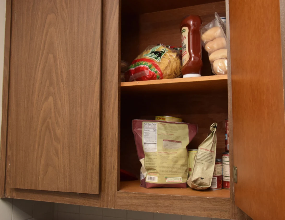 Open pantry cupboard with bread rolls, ketchup, snack bags and canned food.