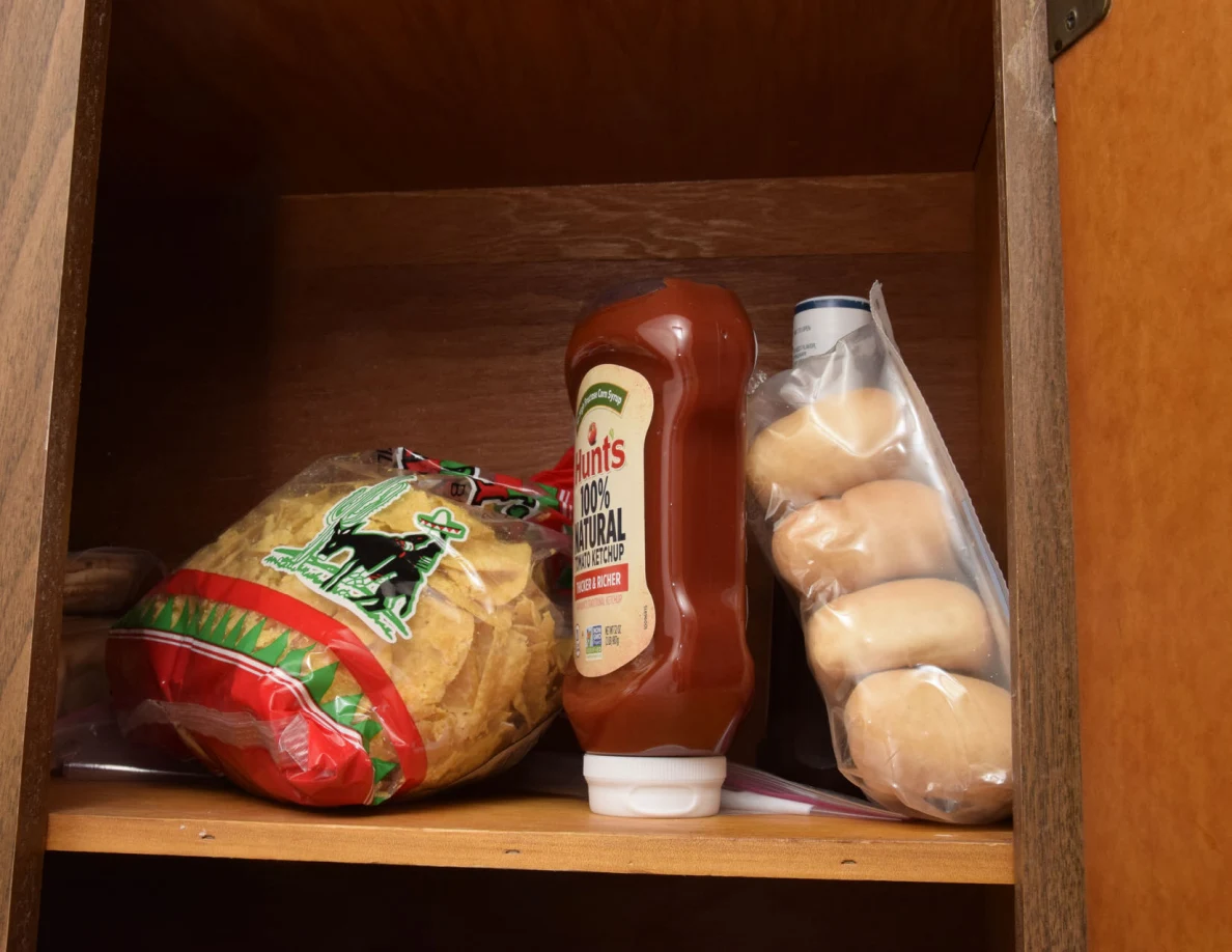 Bag of tortilla chips, ketchup bottle and packaged bread rolls on a pantry shelf.