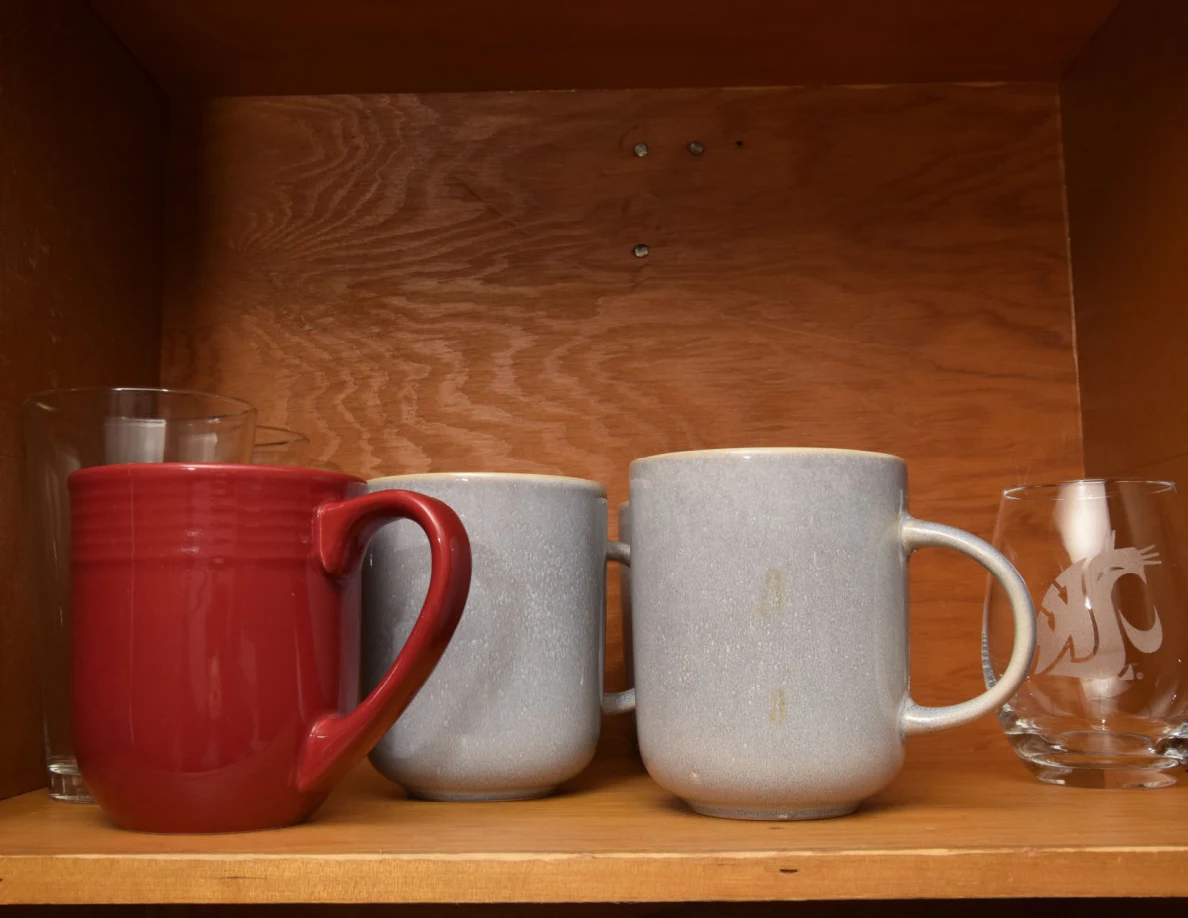 Red and gray mugs and clear drinking glasses lined up on a wooden shelf.