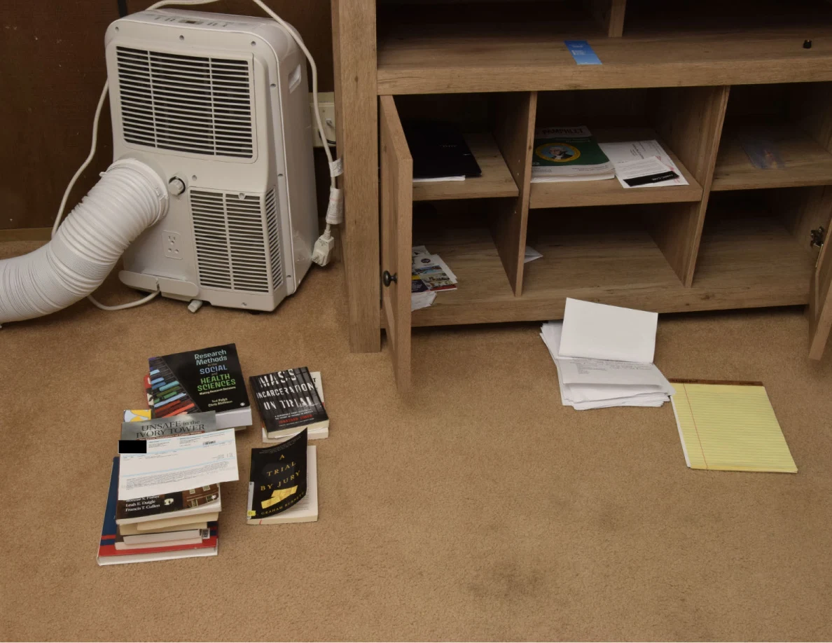 Portable air conditioner beside an open media cabinet, with books, papers and a yellow legal pad on the carpet.