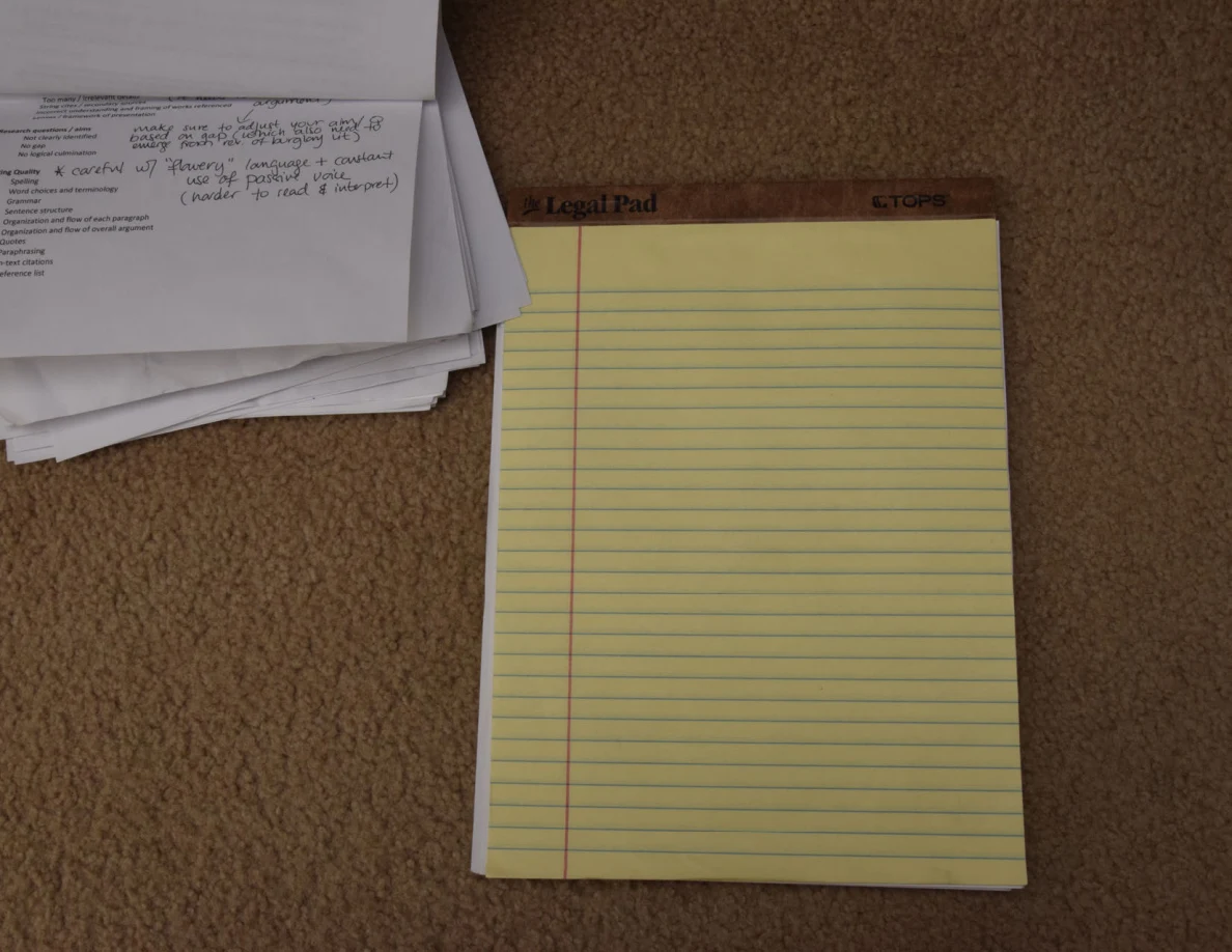 Yellow lined legal pad beside a stack of printed pages with handwritten annotations on carpet.