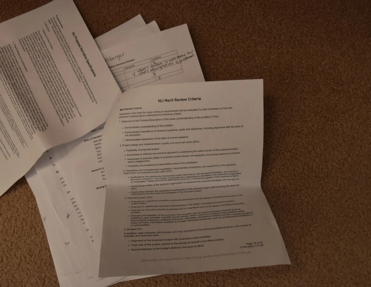 Printed merit-review criteria and other annotated pages spread across carpet.