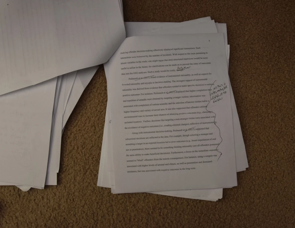 Printed essay page with handwritten notes and a bracket along the right margin, on a paper stack on carpet.