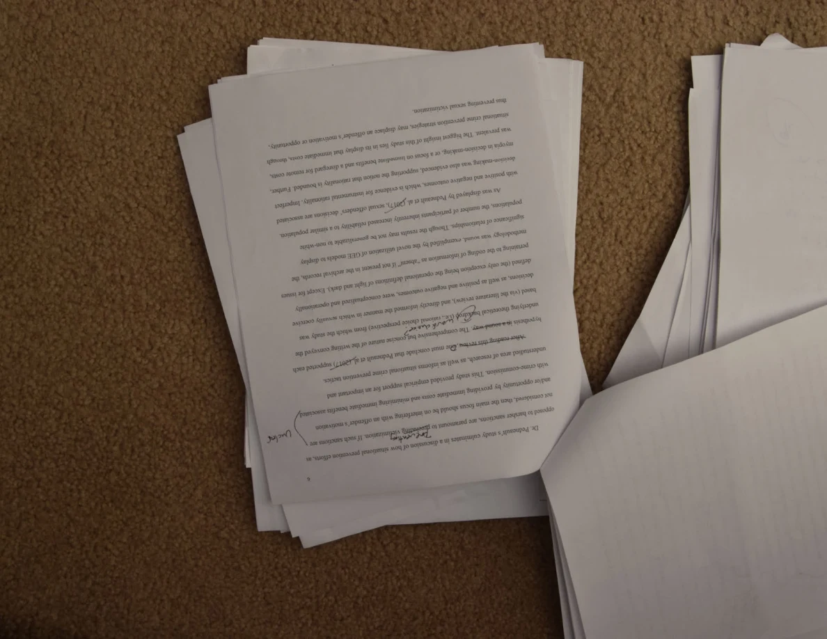 Upside-down printed essay page with handwritten annotations on a stack on carpet.