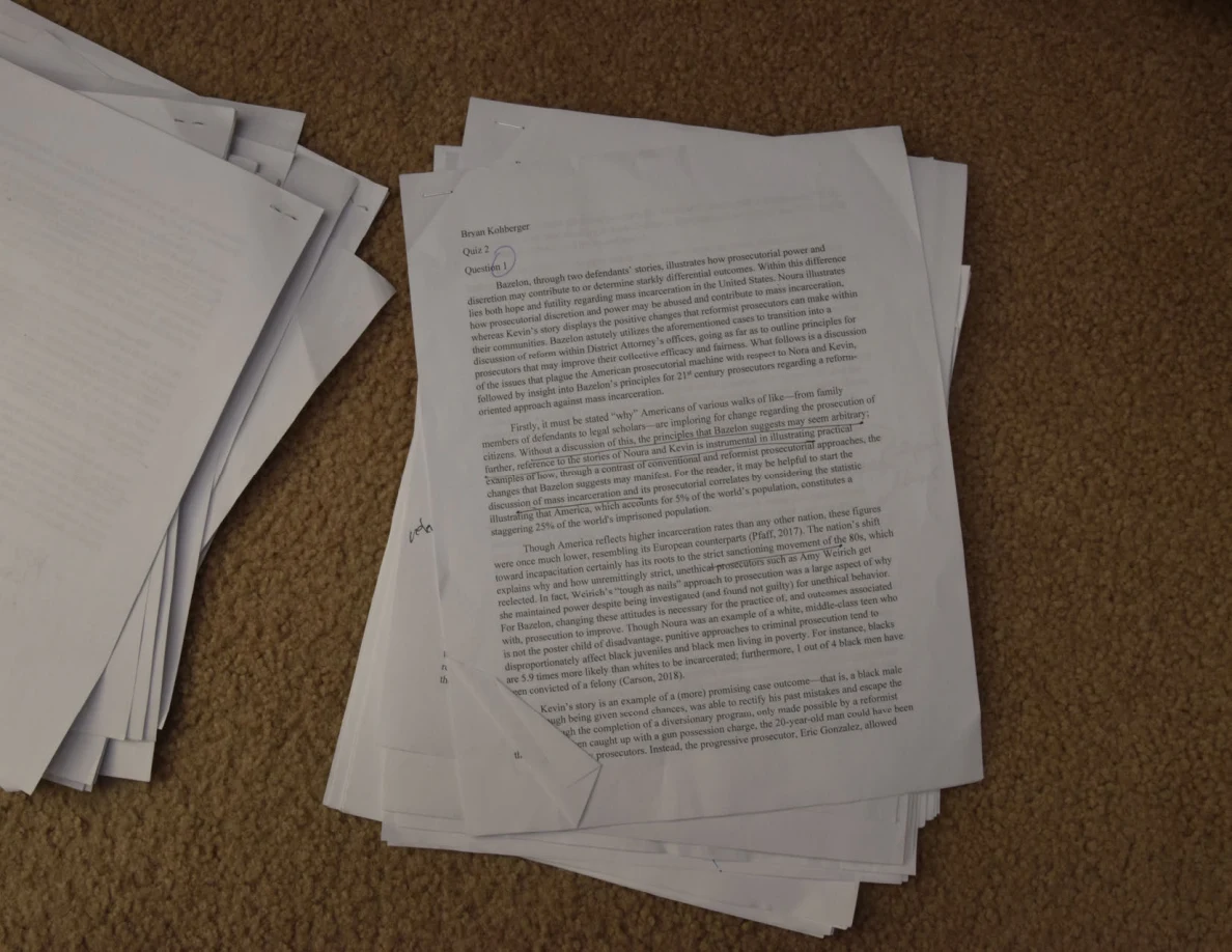 Printed essay page with handwritten marks beside paragraphs, on a stack on carpet.