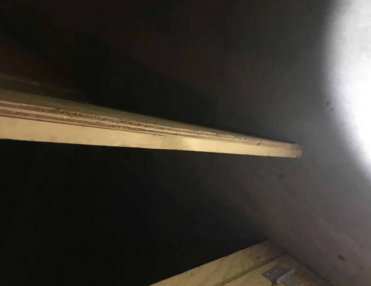 Side view of a plywood panel edge beside a gray wall and dark opening.