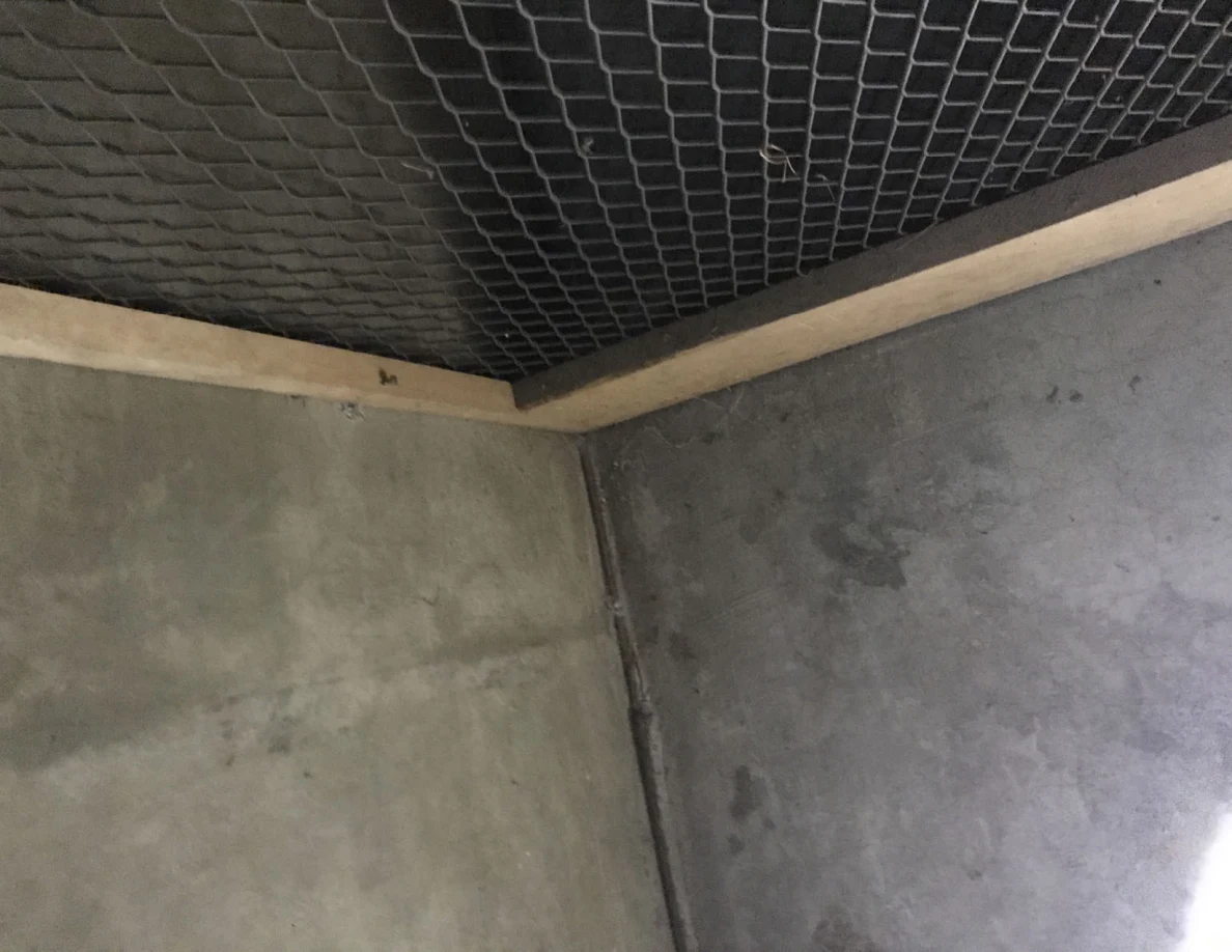 Upper corner of a storage compartment showing mesh, wooden framing and gray walls.