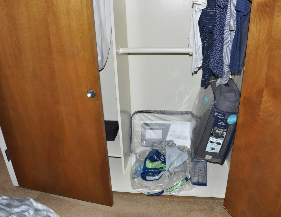 Lower section of an open closet with hanging shirts and packaged bedding on its bottom shelf.