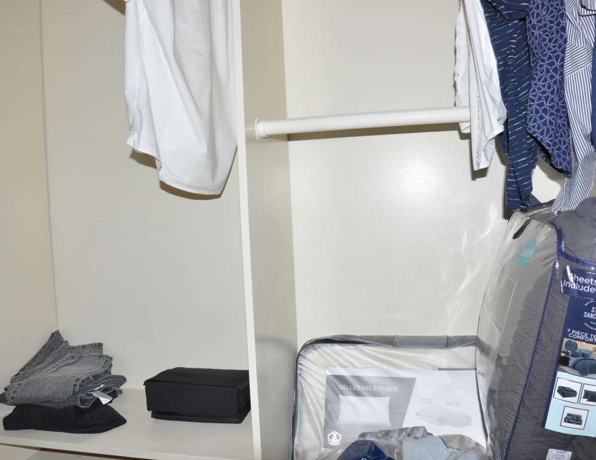 Open closet with hanging shirts, folded clothing, a black case and packaged bedding.