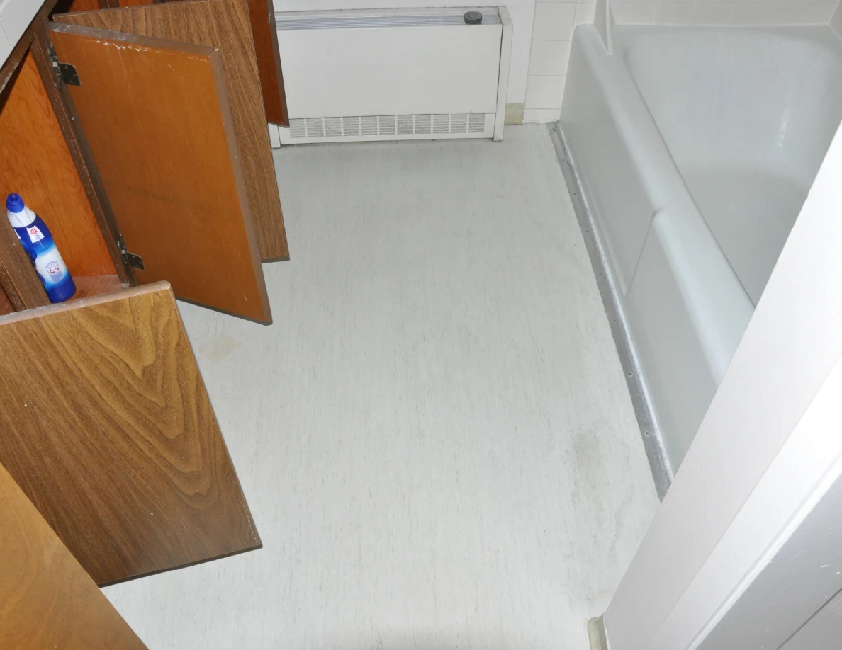 Bathroom floor between an open wooden vanity cabinet and white bathtub, with wall heater beyond.